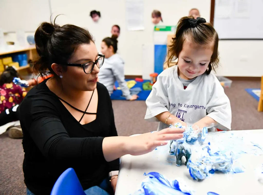 A teacher and a young girl in kindergarten play with blue shaving cream on a desk.