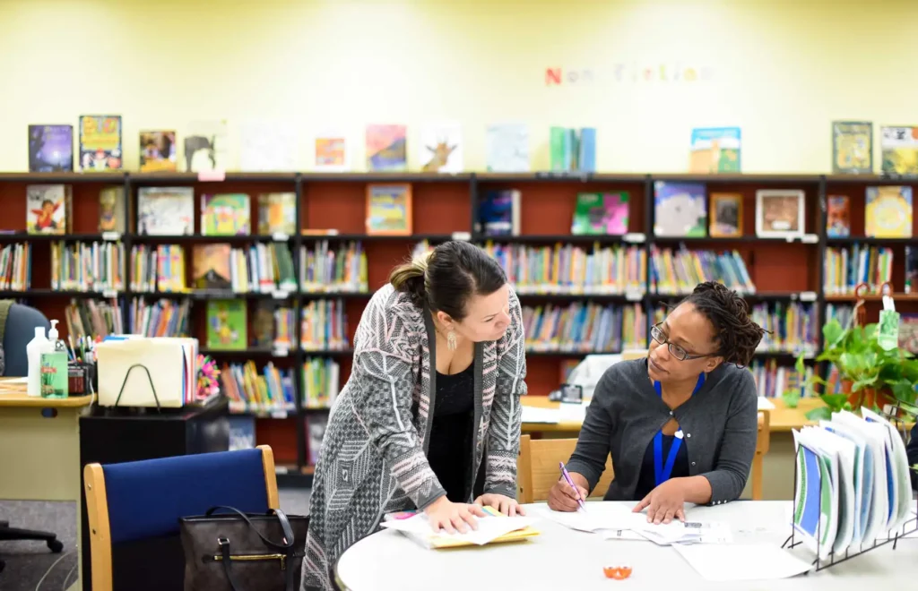 Two teachers discuss paperwork in a school library.