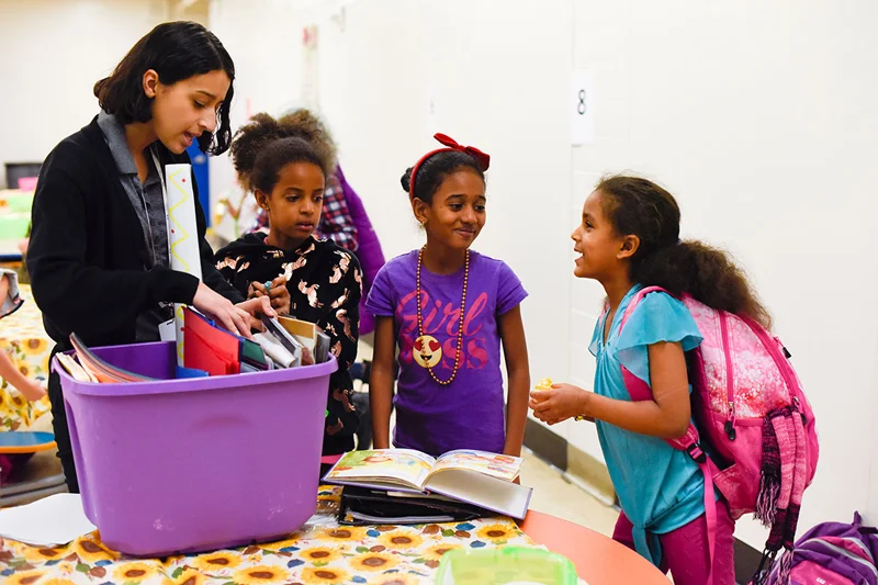 A group of young girls wearing brightly colored shirts and their teacher look through a container full of notebooks.