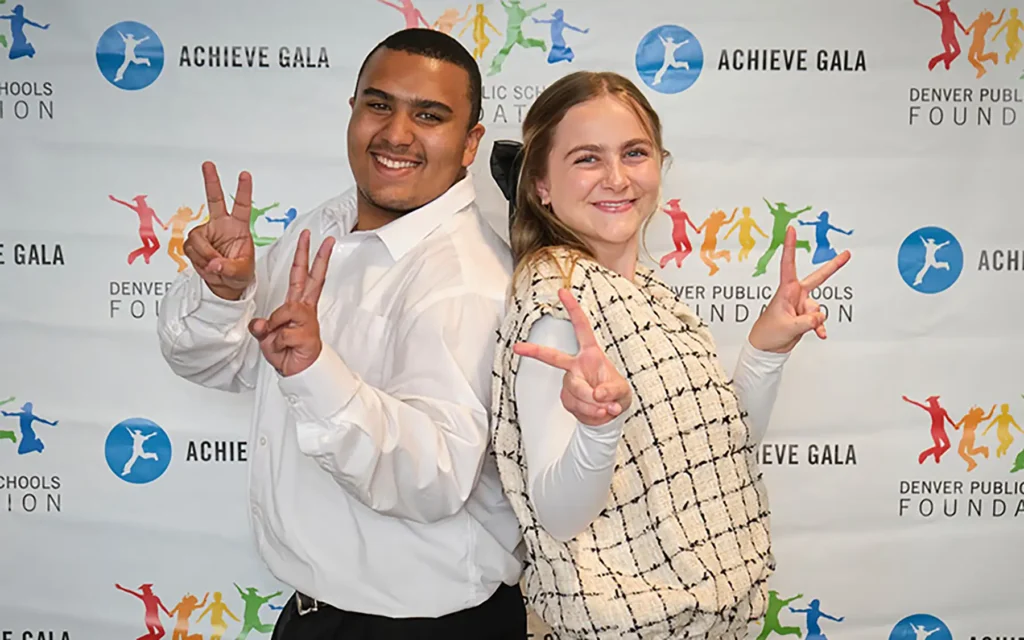 Two individuals pose with peace signs in front of a backdrop with "Achieve Gala" and "DPS Foundation" logos printed across it.