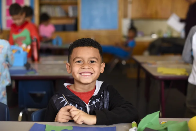 A little boy sits at his desk in a classroom during craft time and smiles.