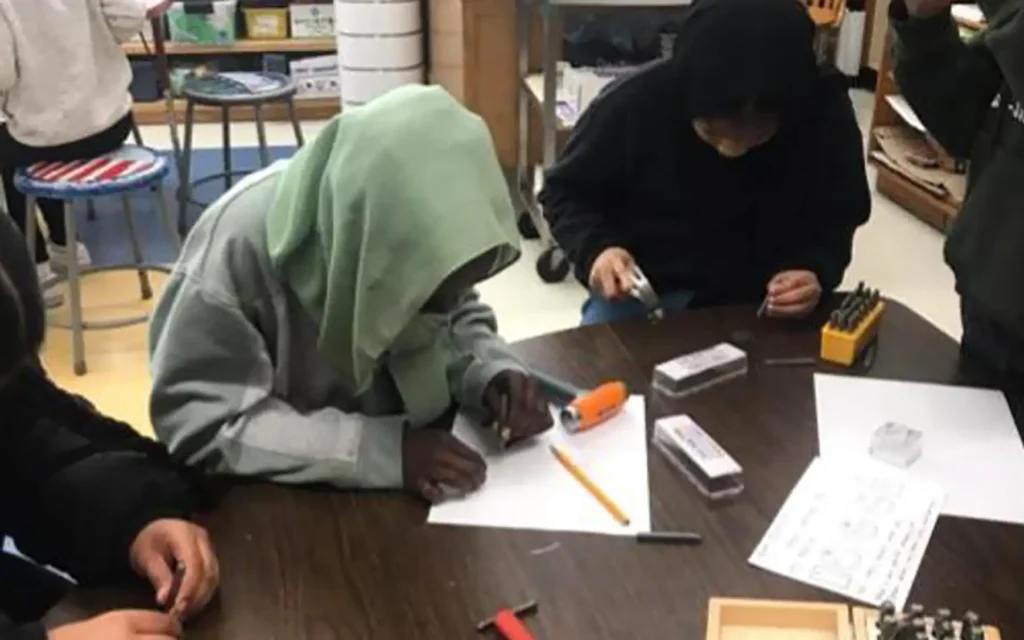 Students at a table look down at pieces of paper and tools as they complete a workshop project.