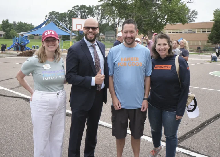 A group of volunteers stand at a school playground while other volunteers perform maintenance behind them.