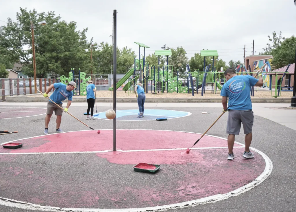 A group of volunteers paint the pavement at a tetherball court at a school.