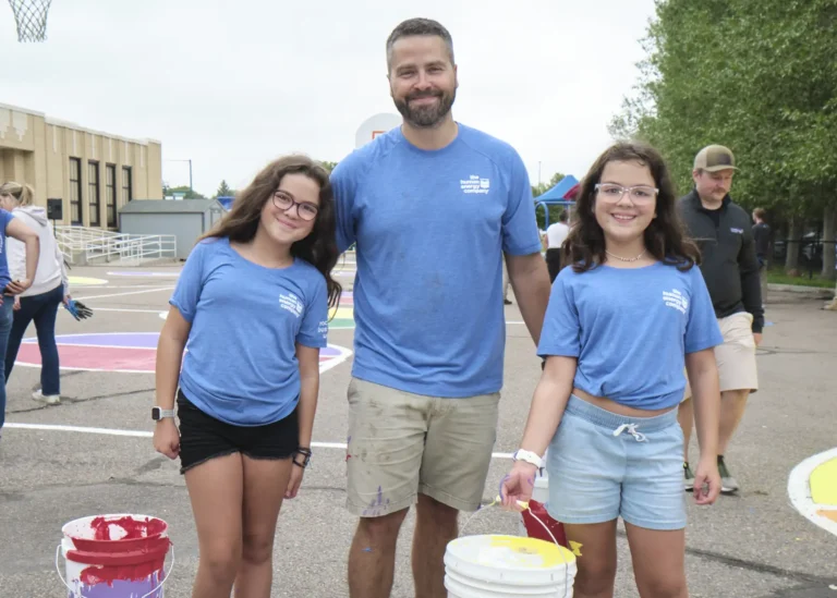 A white man and two young white girls stand in a paved playground outside of a school holding buckets of paint.