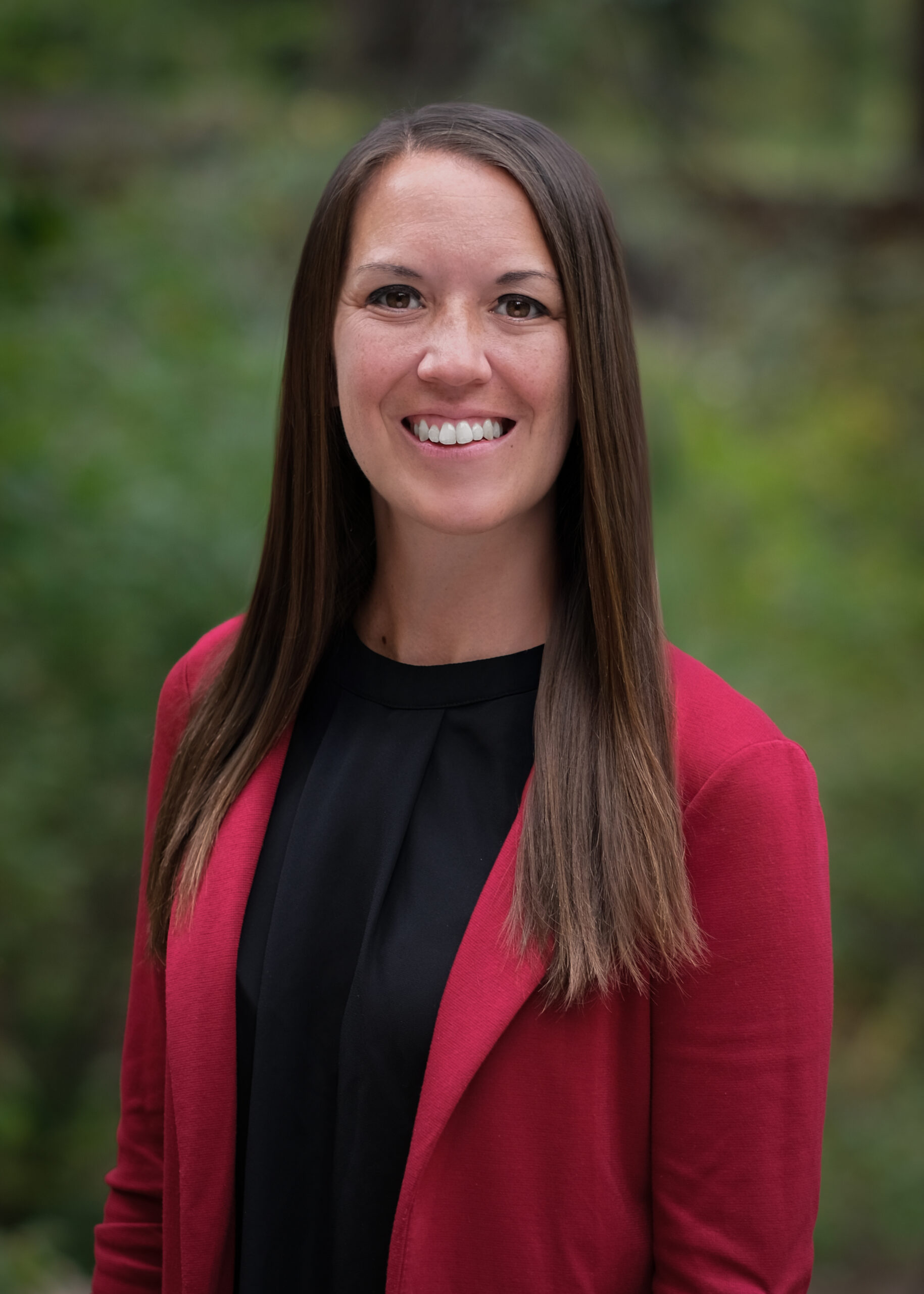 Headshot of Samantha, smiling outdoors in front of greenery, wearing a red blazer over a black top.
