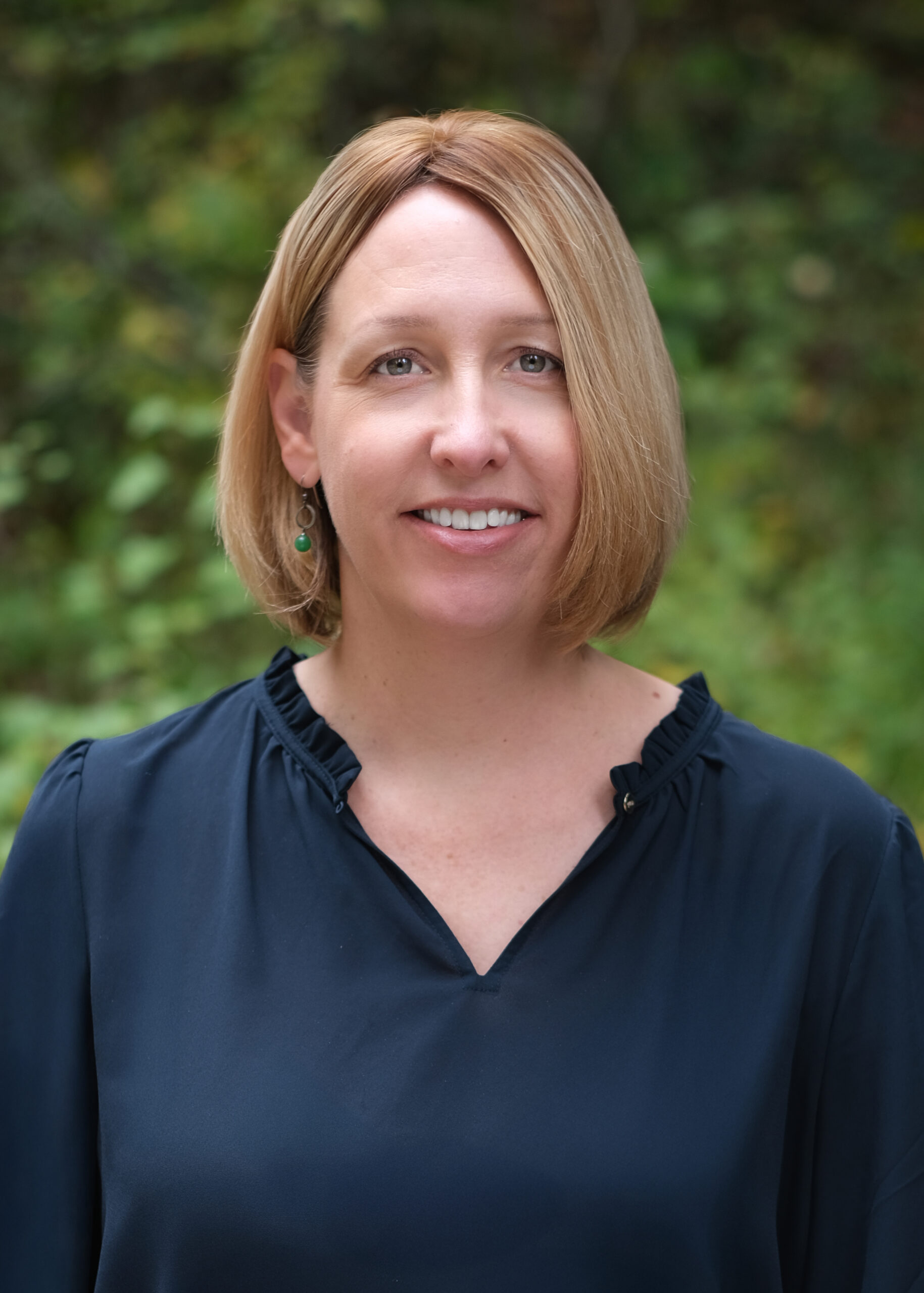 Headshot of Jennifer smiling outdoors in front of greenery, wearing a navy blouse with ruffle detail at the collar.