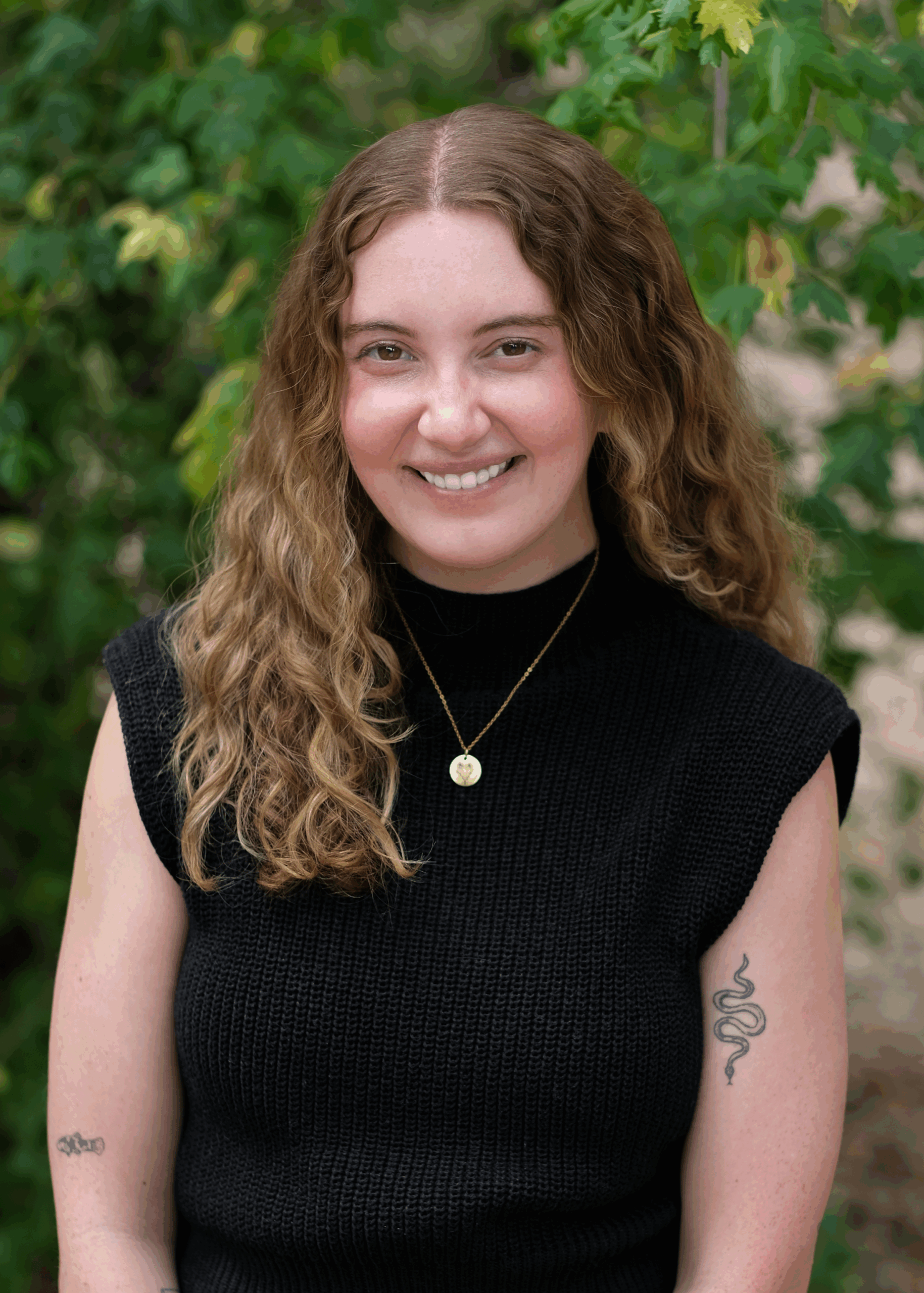 Headshot of Erin, smiling outdoors in front of greenery, wearing a black sleeveless sweater and gold necklace.