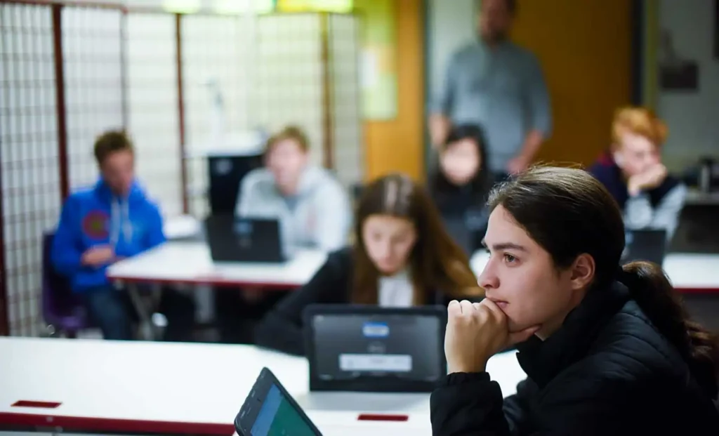 A student sits at a table with a laptop in a classroom with other students.