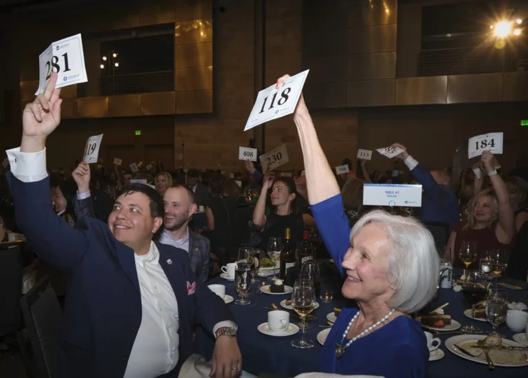 Donors fill a large room during an auction at the Achieve Gala, a large amount of them raising their paddles to make a donation.