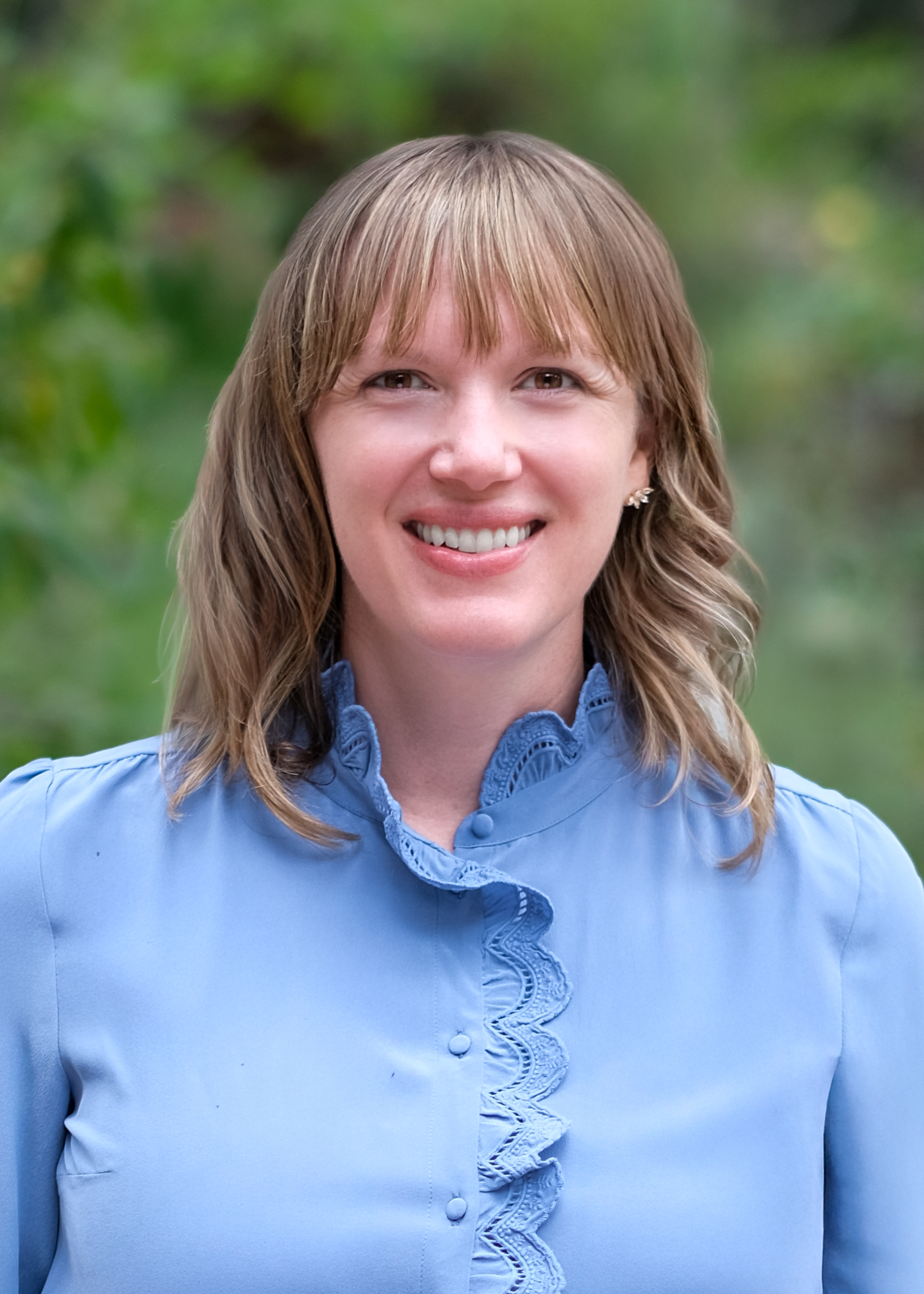 Headshot of Sara smiling outdoors in front of greenery, wearing a light blue blouse with lace detailing.