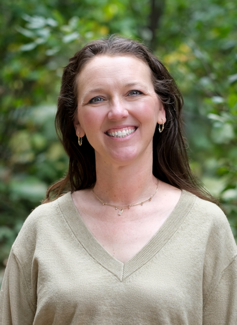 Headshot of Jen smiling outdoors in front of green foliage, wearing a tan sweater and gold jewelry.