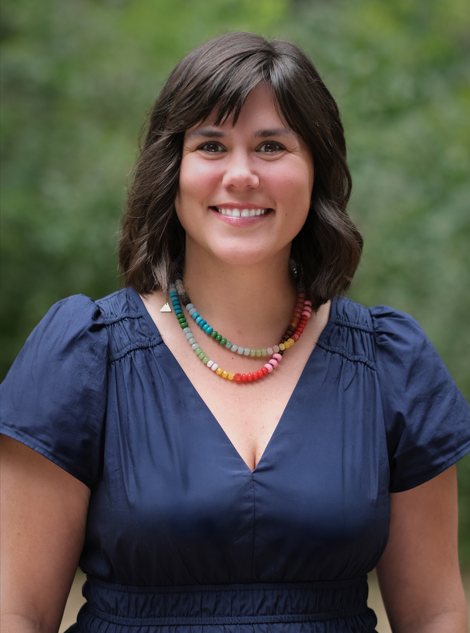 Headshot of Mary, smiling outdoors in front of greenery, wearing a navy blue blouse and a colorful beaded necklace.