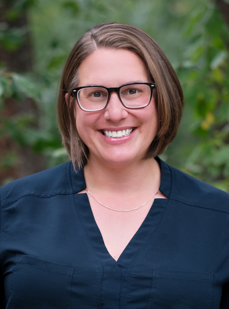 Headshot of Lauren, smiling outdoors in front of greenery, wearing a navy blue blouse.
