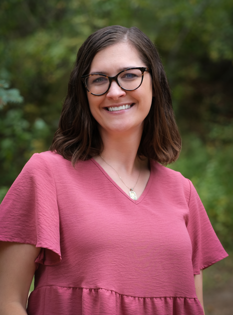 Headshot of Ashley smiling outdoors in front of greenery, wearing glasses and a rose-colored blouse.