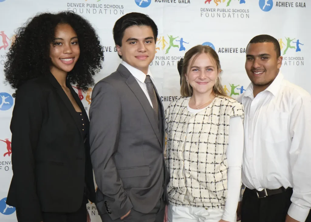 Four students of mixed races in formal attire stand in front of a backdrop that has DPS Foundation logos and Achieve Gala logos.