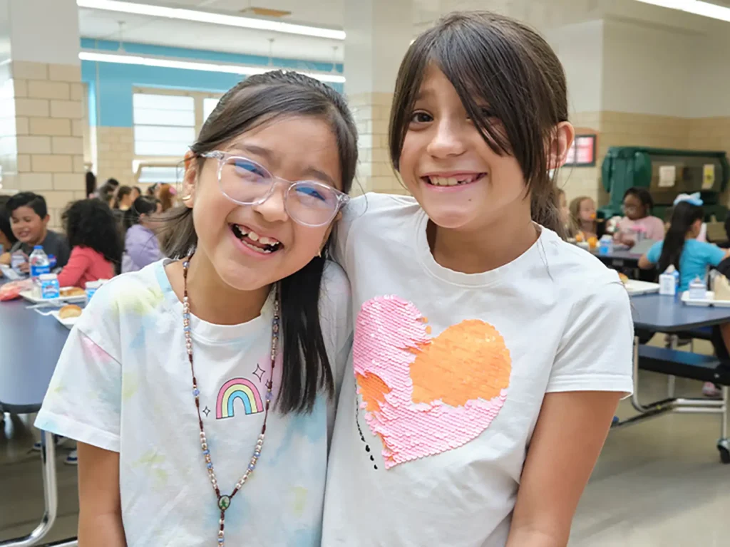 Two elementary aged girls laugh together in their school cafeteria.