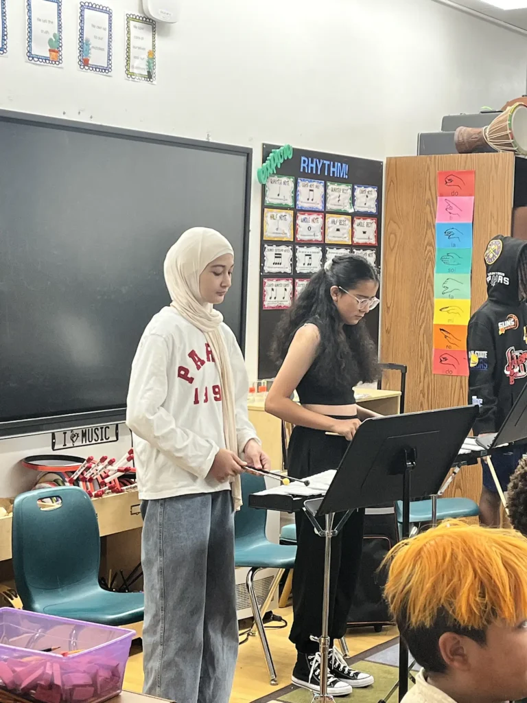 A hijabi student and a student with curly dark brown hair stand at a xylophone, hitting the keys with mallets.