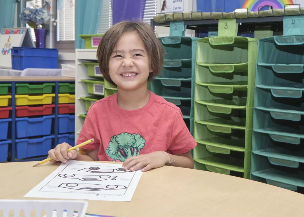 An elementary aged student sits at a table working on a worksheet with three car graphics on it.