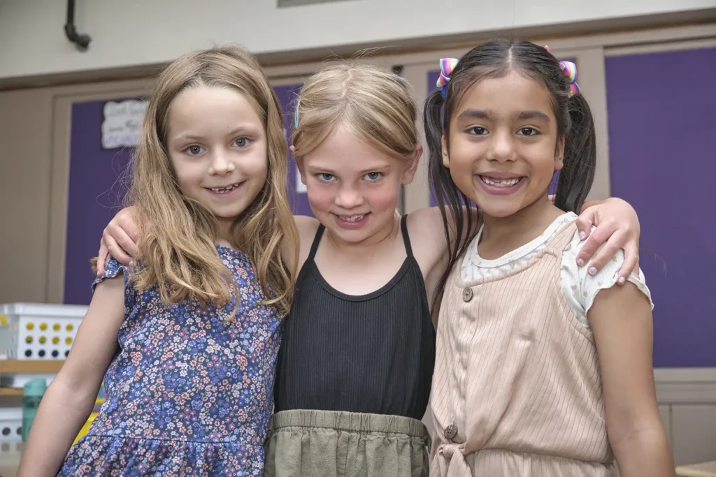 Three elementary aged girls smile together in their classroom.