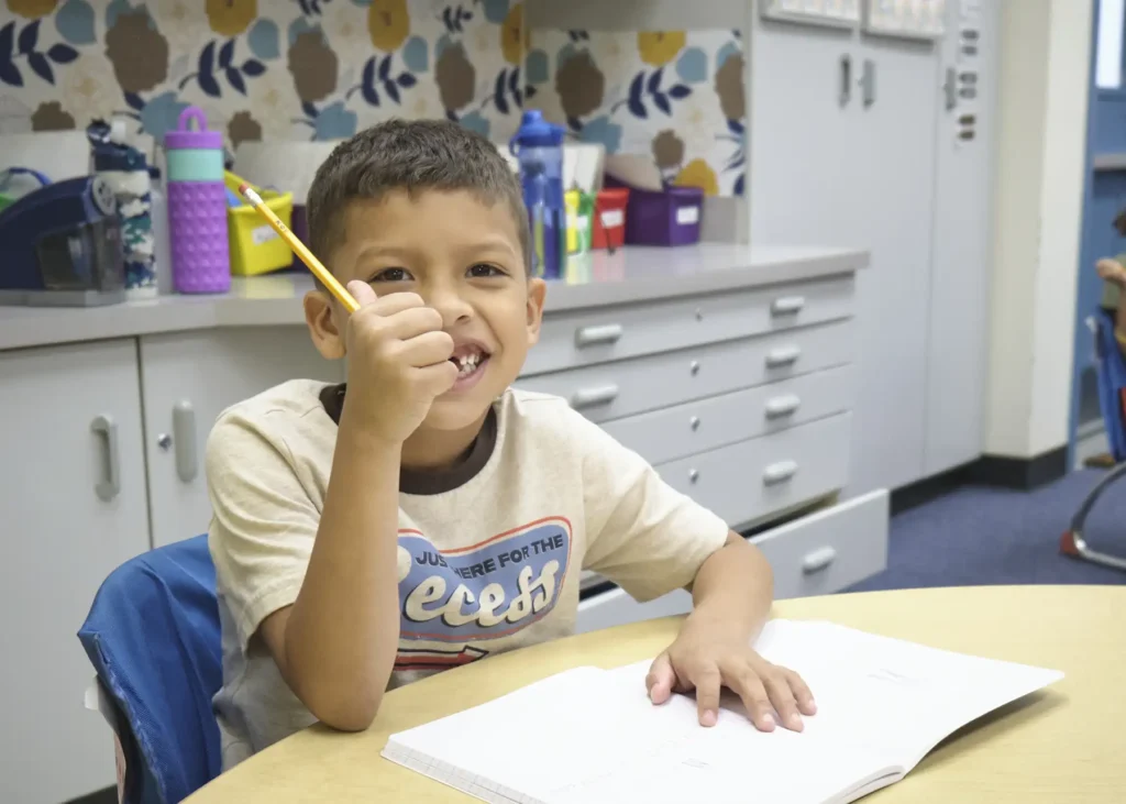 A young boy sits at a desk with a pencil in his hand and works in a journal.
