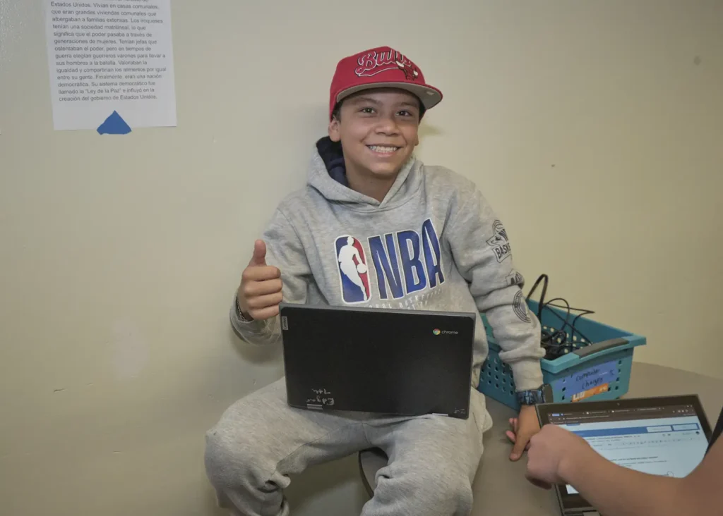 A student wearing an NBA sweatshirt sits at a table with a Chromebook in his lap.