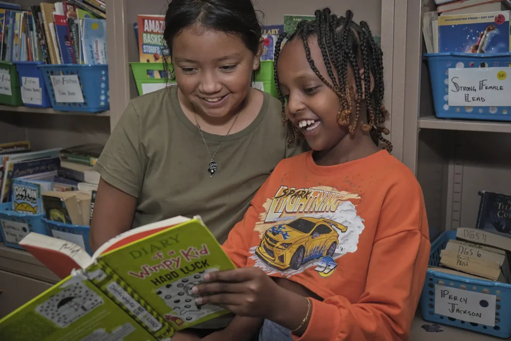 Two elementary aged students of color reading "Diary of a Wimpy Kid: Hard Luck" in their classroom.