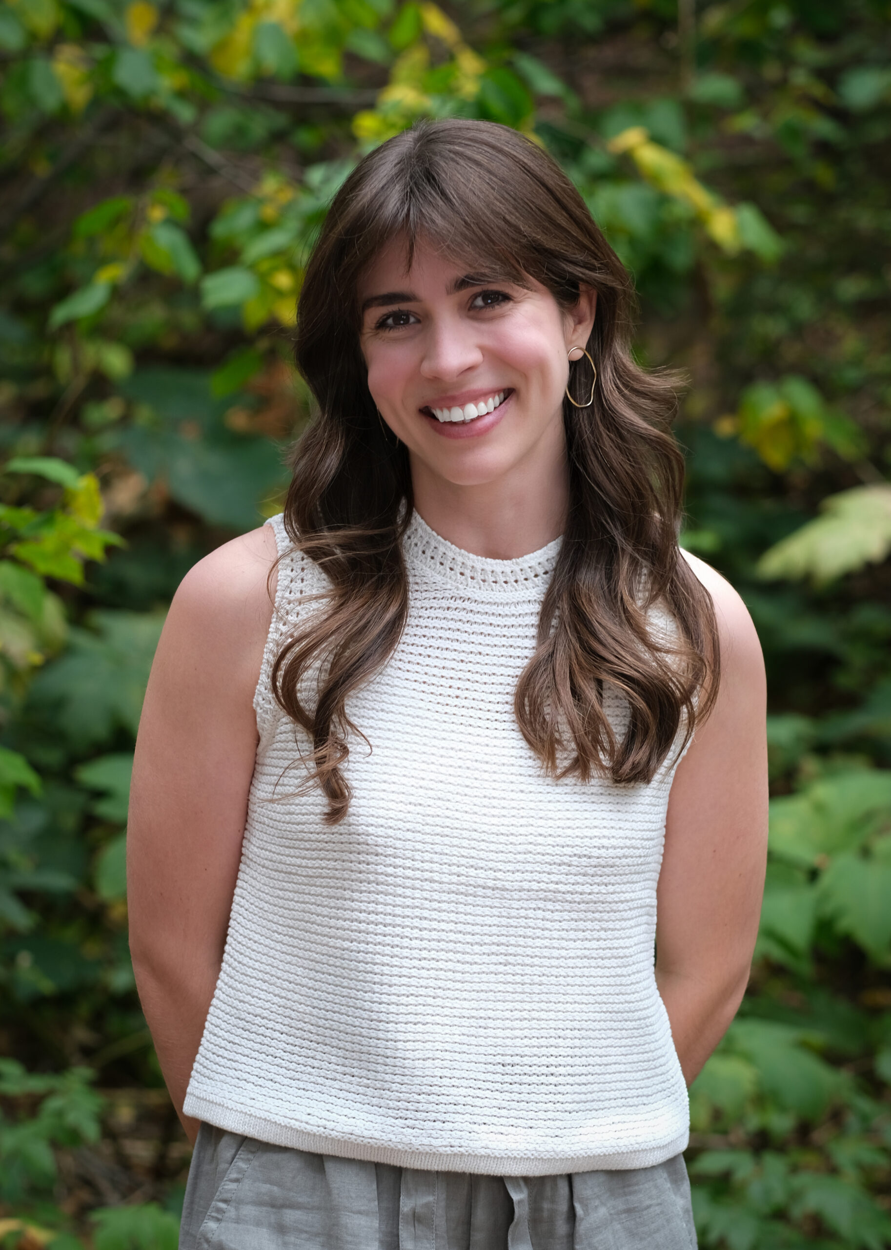 Headshot of Valeria, smiling outdoors in front of greenery, wearing a white sleeveless knit top and gray pants.