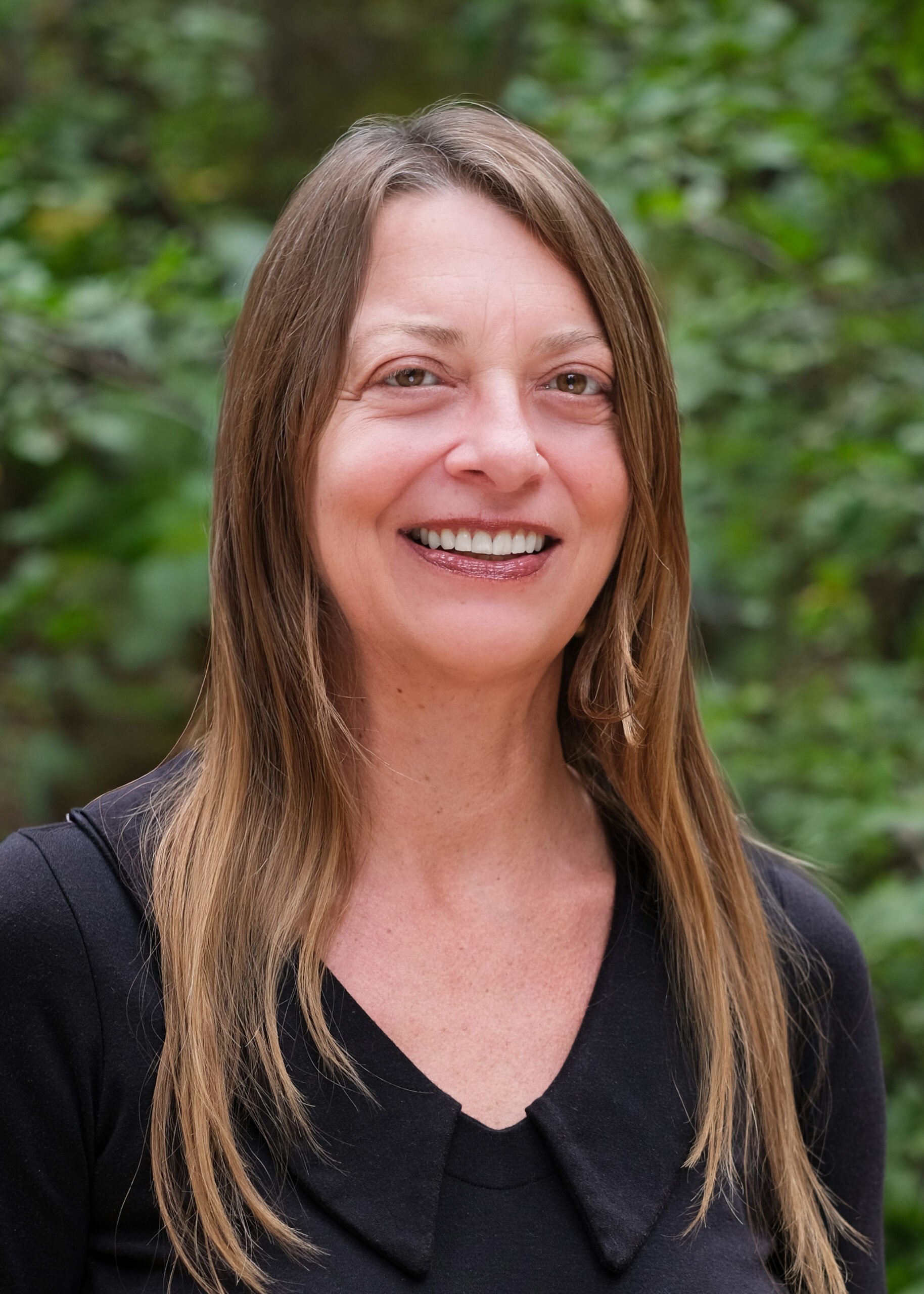 Headshot of Crystal, smiling outdoors in front of greenery, wearing a black top with long light brown hair.