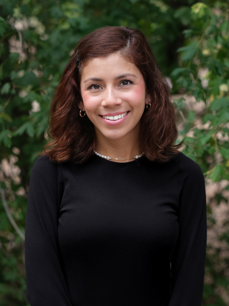 Headshot of Angelica, smiling outdoors in front of greenery, wearing a black long-sleeve top.