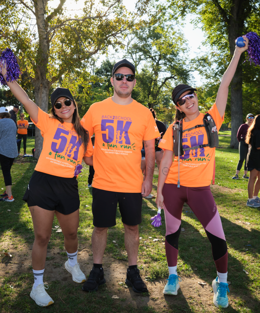 Three participants pose together at the Back to School 5K Fun Run, smiling and raising pom-poms in celebration.