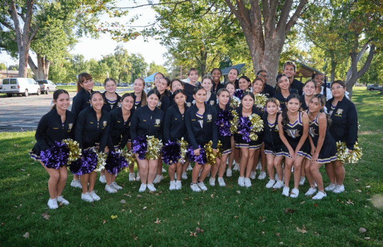 A cheer team poses together outdoors, smiling and holding purple and gold pom-poms.