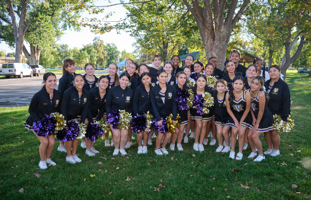 A cheer team poses together outdoors, smiling and holding purple and gold pom-poms.