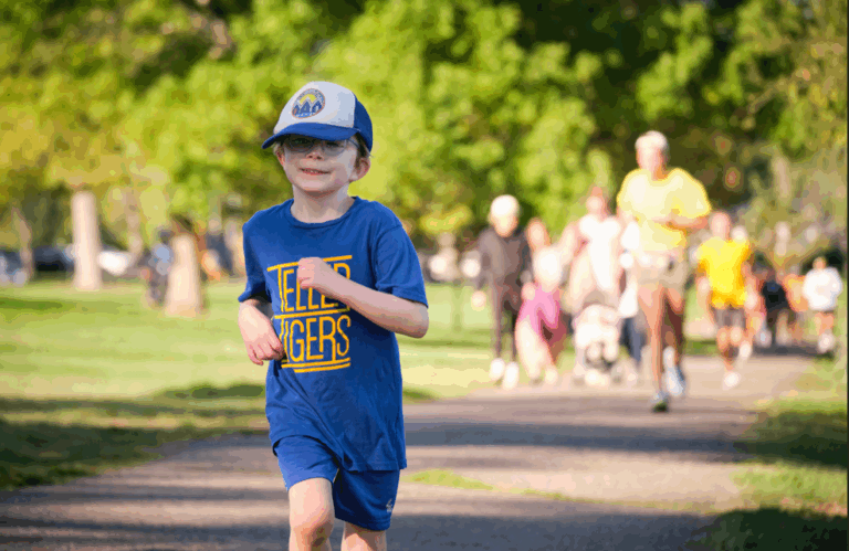 A young student wearing a blue shirt and hat runs on a path with other participants in the background.