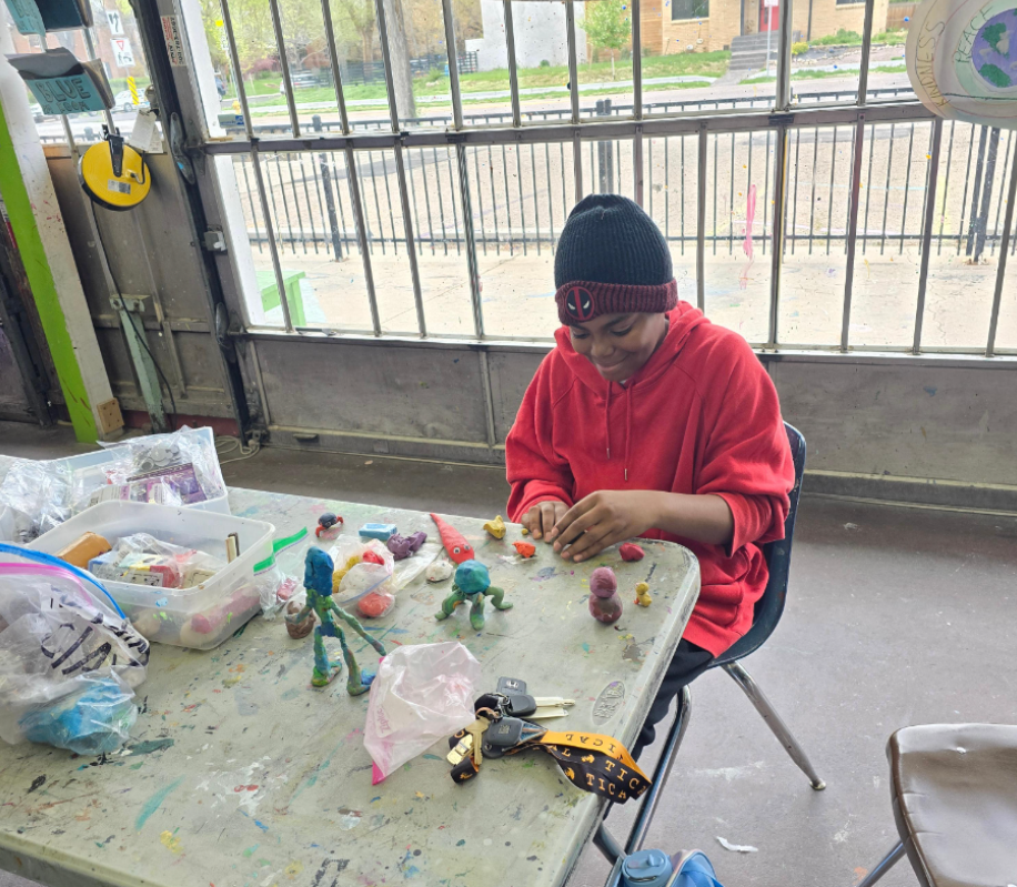 A student wearing a red hoodie and black beanie sits at a table shaping colorful clay figures.