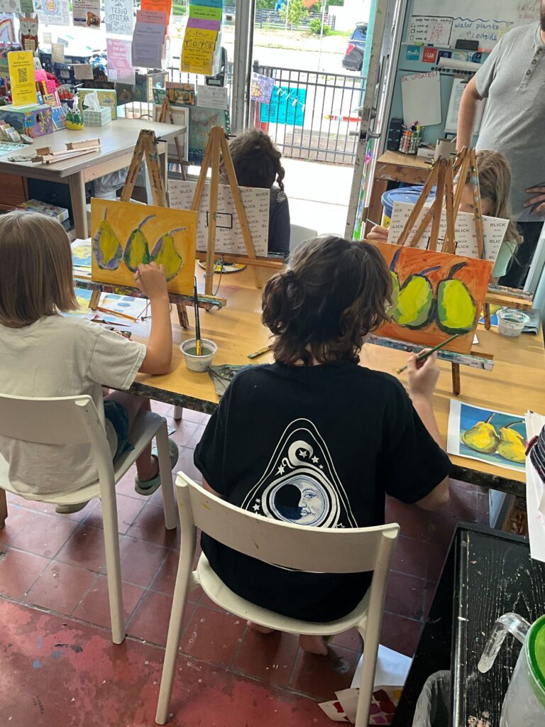 Children seated at tables painting on small canvas easels in an art classroom.