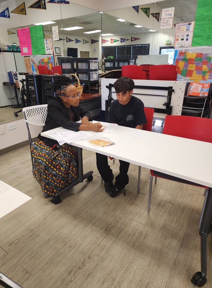 A teacher sitting with a student at a table reviewing schoolwork in a classroom.