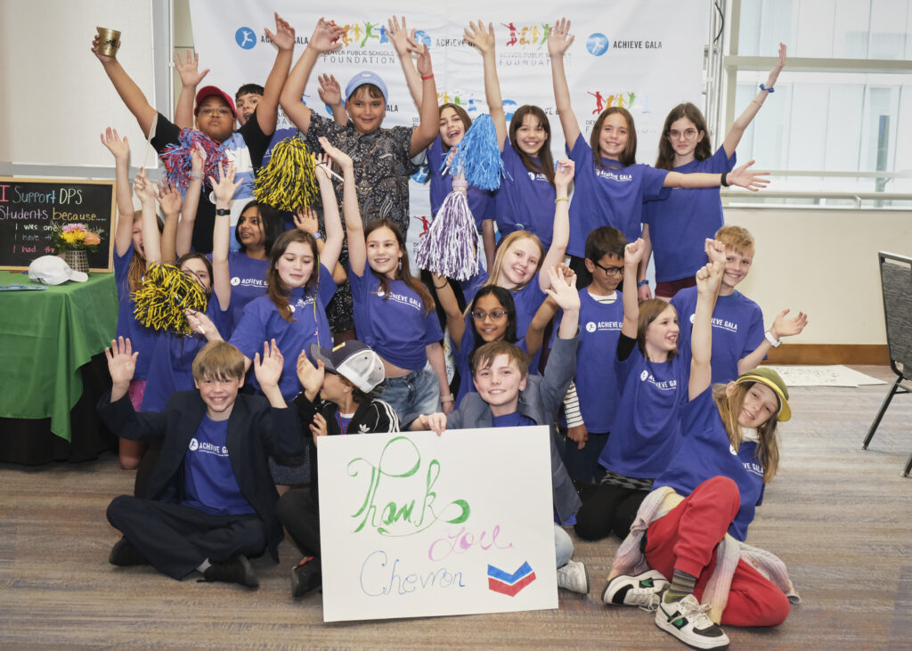 Group of students in blue shirts smiling and cheering with raised hands with a sponsor sign in the front.