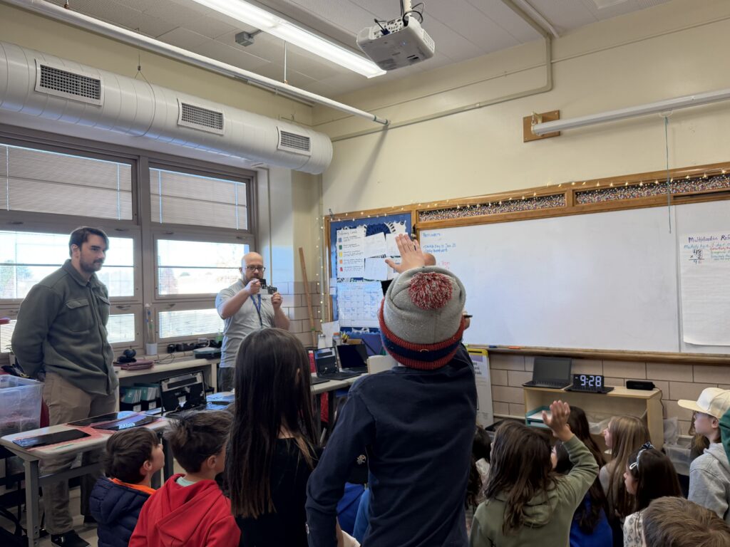 Students raise their hands while a teacher demonstrates something at the front of a classroom.