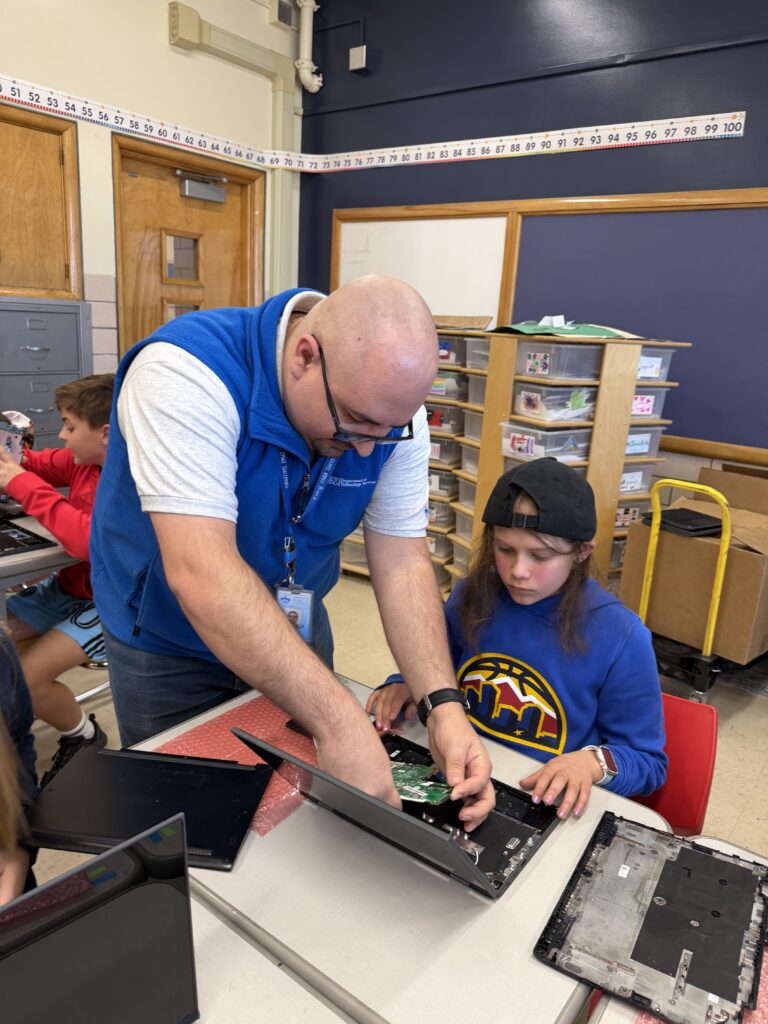 A teacher helps a student repair the inside of a laptop at a classroom desk.