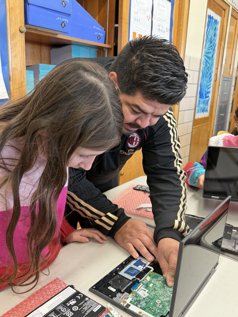 A teacher helps a student repair the inside of a laptop at a classroom desk.