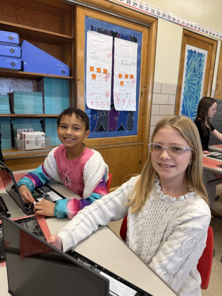 Two students smile while working on an open laptop in a classroom.