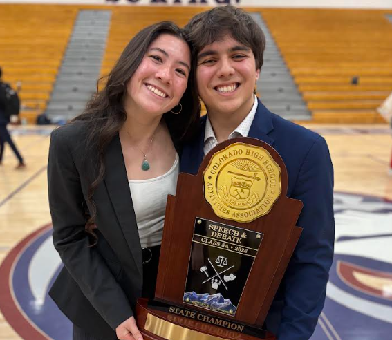 Two students in formal attire smiling and holding a state champion speech and debate trophy in a gymnasium.