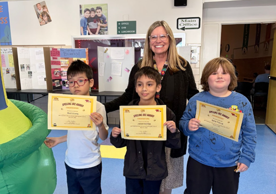 Three elementary school students holding award certificates stand with a smiling adult in a school hallway.